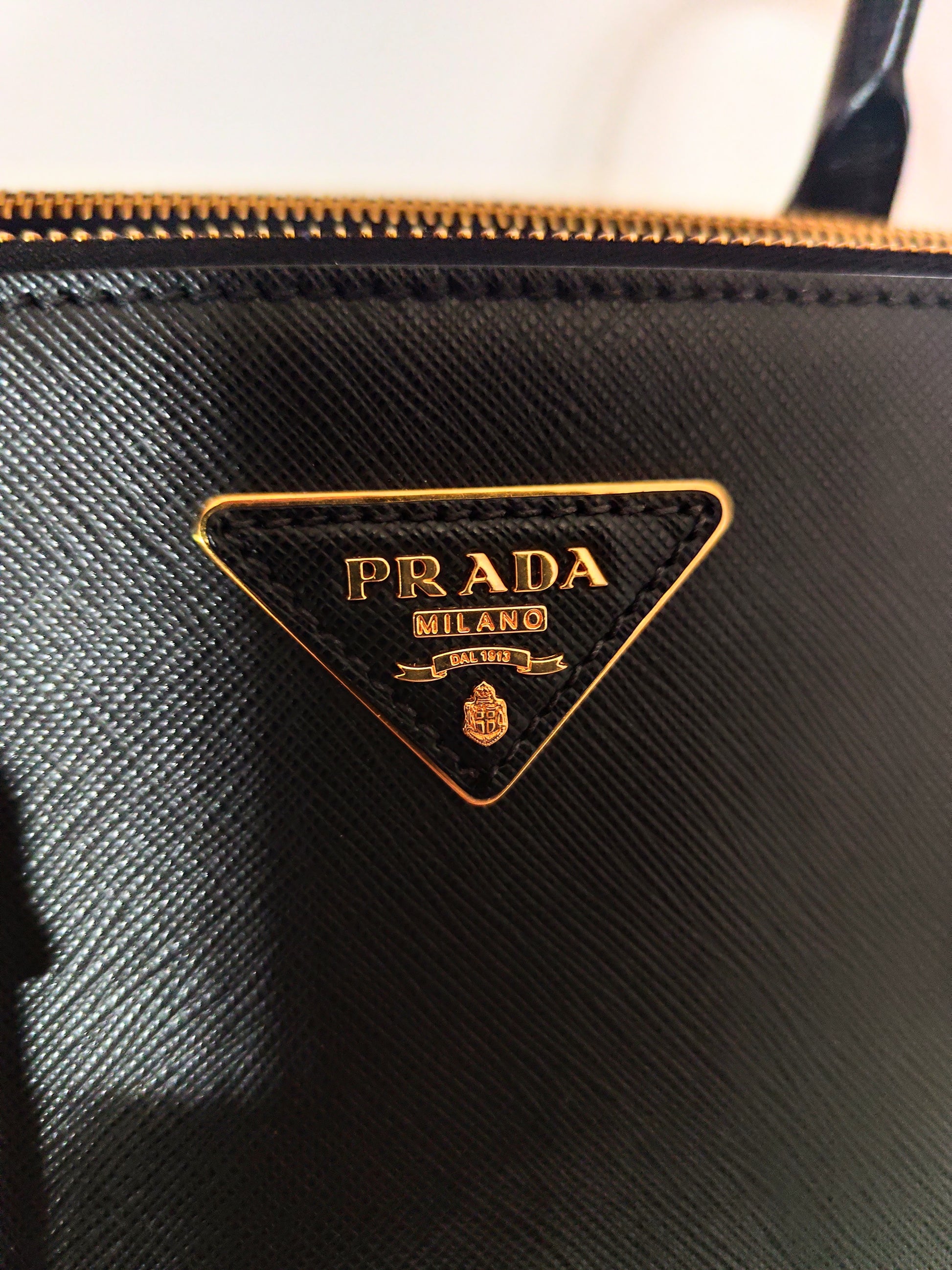 Dettaglio ravvicinato del logo Prada dorato su pelle nera saffiano, con zip in metallo visibile