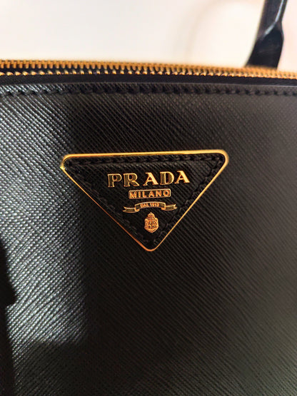 Dettaglio ravvicinato del logo Prada dorato su pelle nera saffiano, con zip in metallo visibile